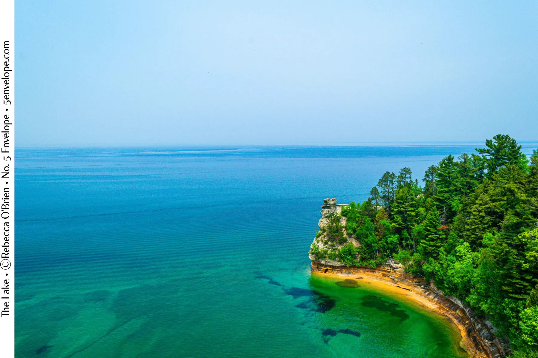 Photograph titled The Lake by Rebecca O'Brien: an aerial view of a forested rock point meeting turquoise water.